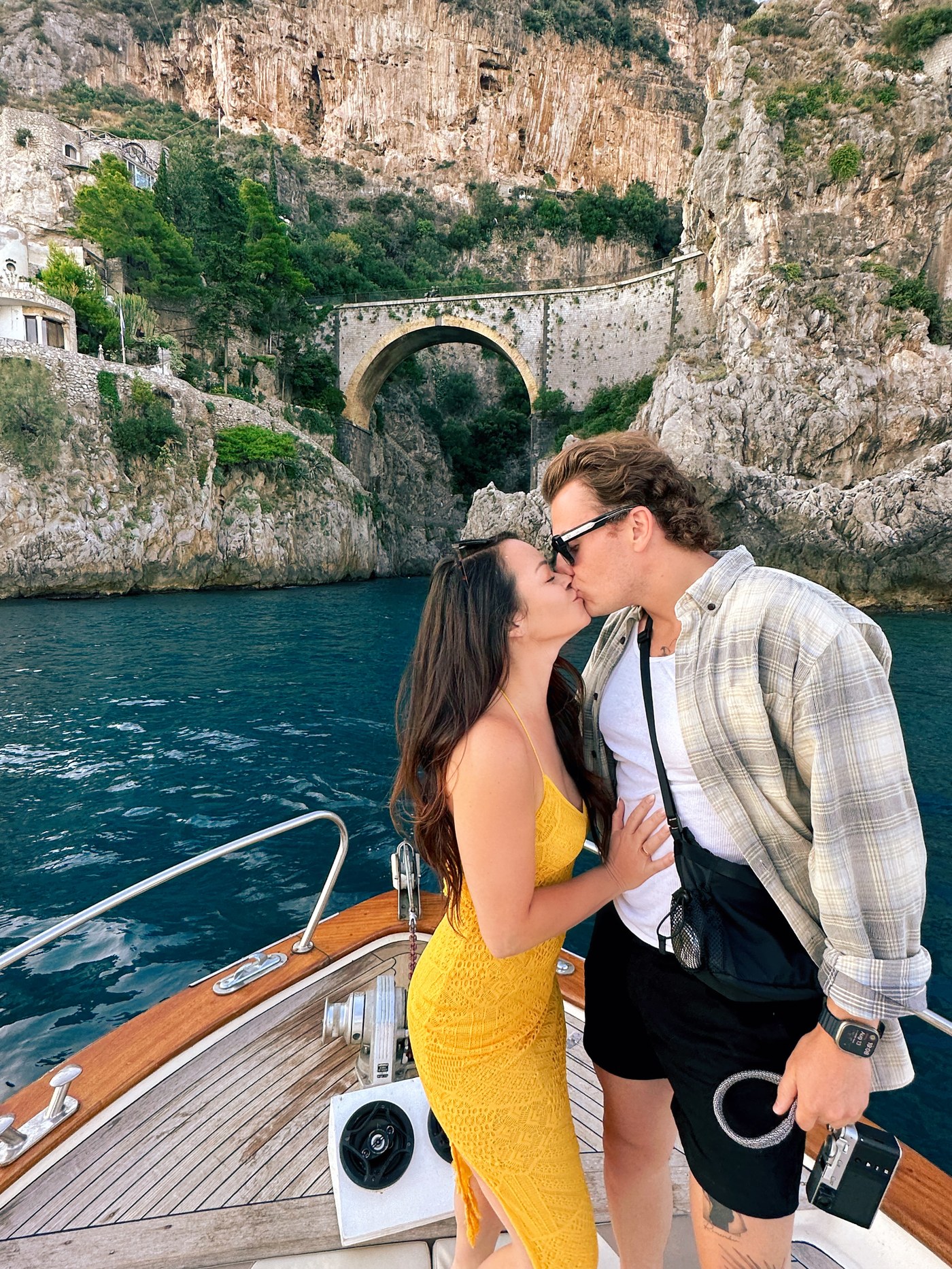 Sean and Brittany kissing on the bow of a boat beneath a stone arch bridge and cliffs