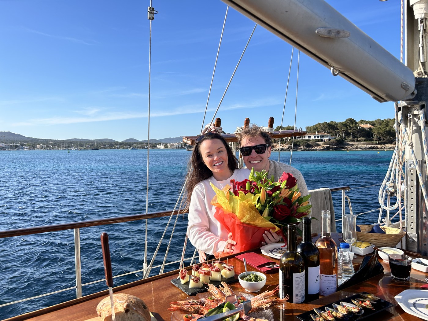 Sean and Brittany on the deck of a sailboat with flowers and a spread of food, coastline behind them