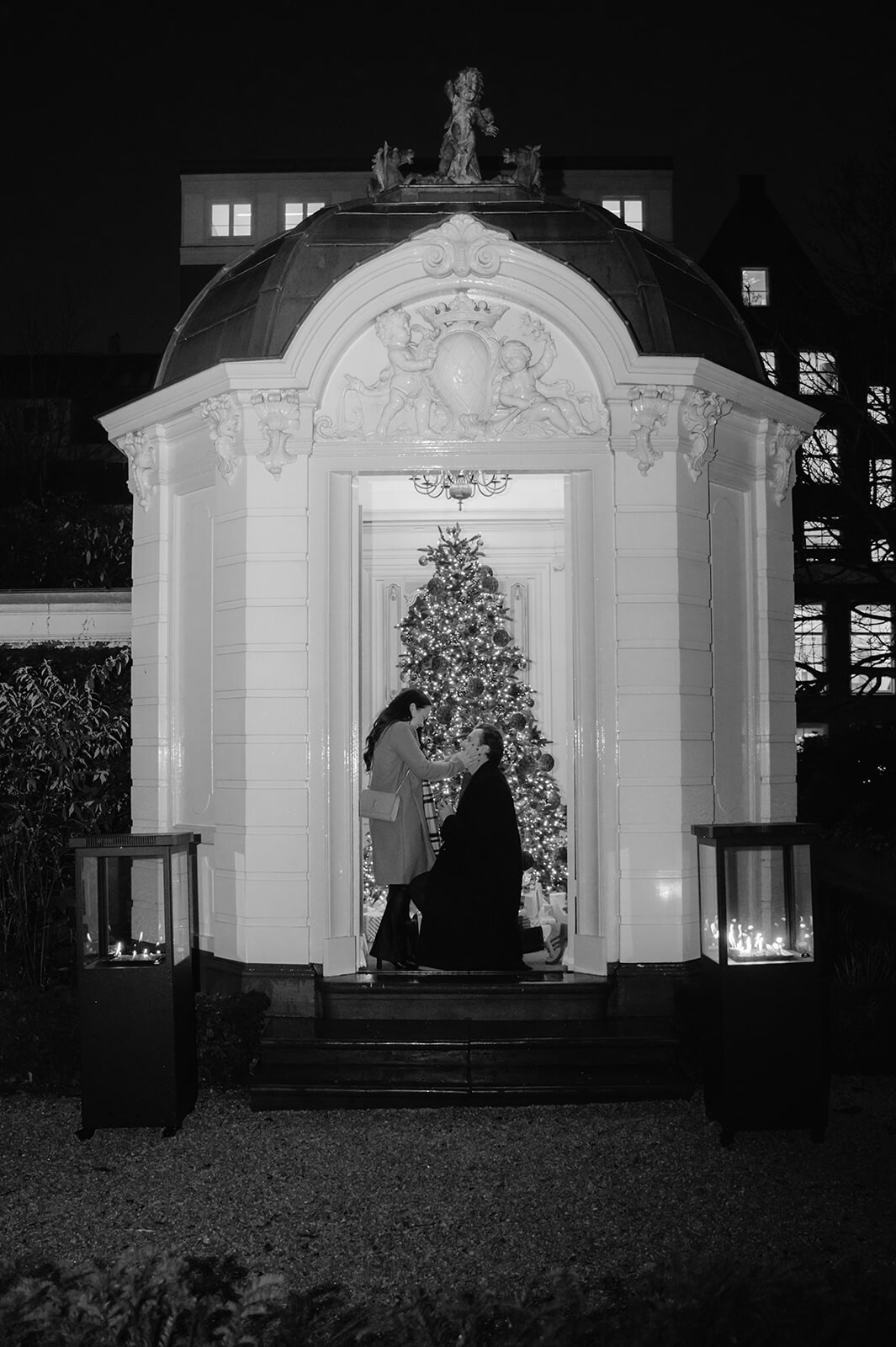 Sean kneeling to propose to Brittany in a candlelit garden pavilion with a Christmas tree glowing behind them