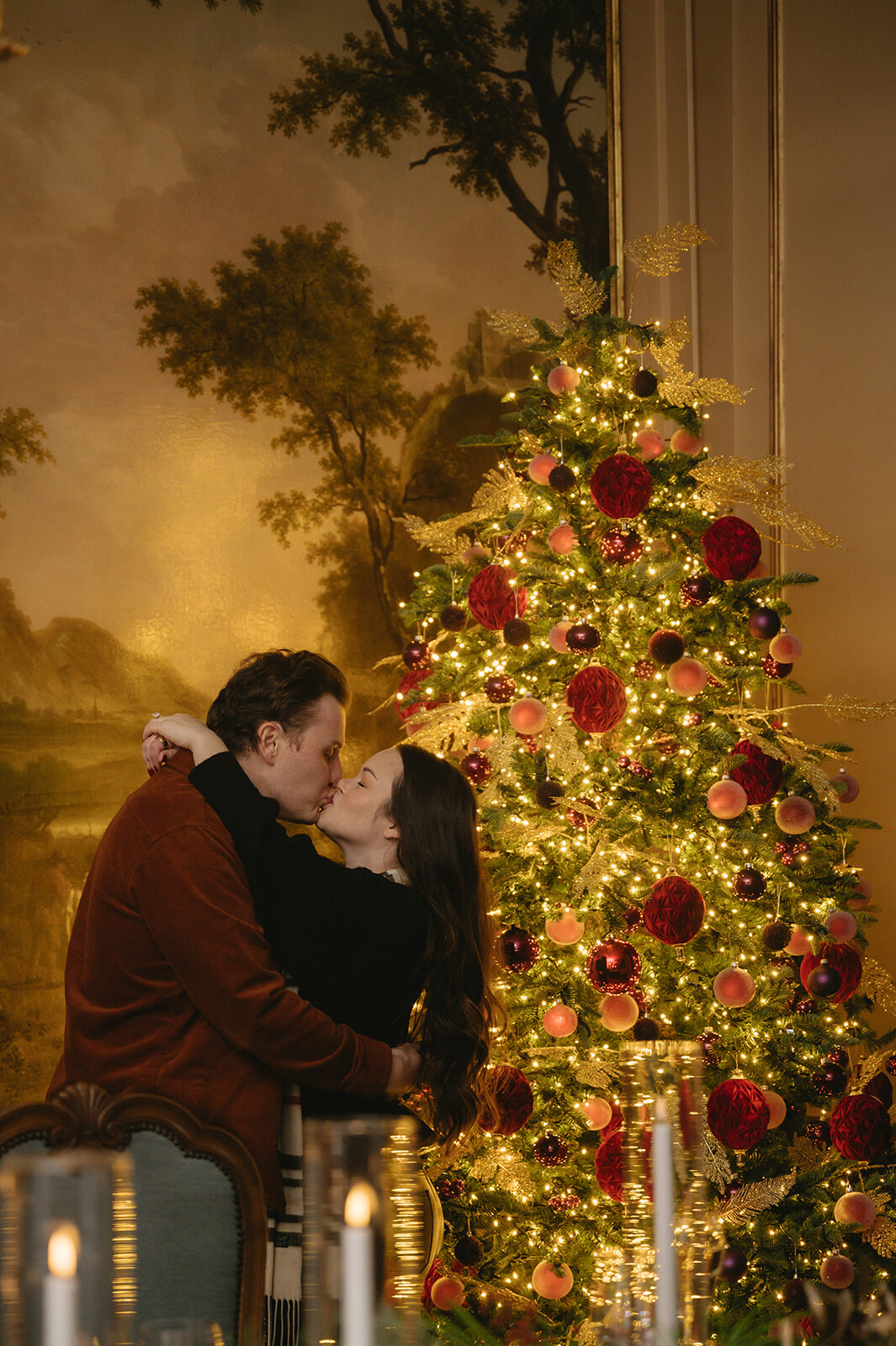 Sean and Brittany kissing beside a Christmas tree dressed in burgundy and gold, her new ring catching the light